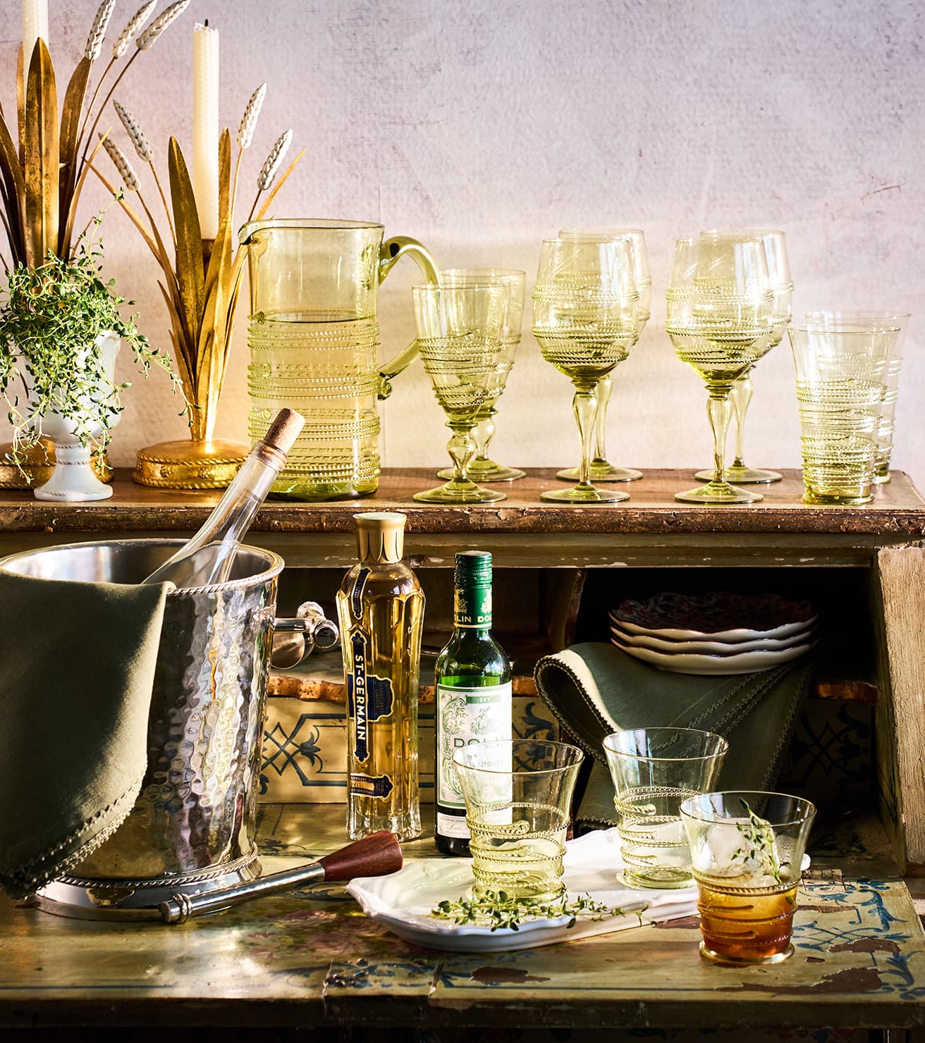 A styled tabletop scene with a set of translucent pale green drinking glasses and a matching glass pitcher on a wooden shelf, alongside a hammered metal ice bucket, bottles, plates, and a glass of amber liquid in the foreground.