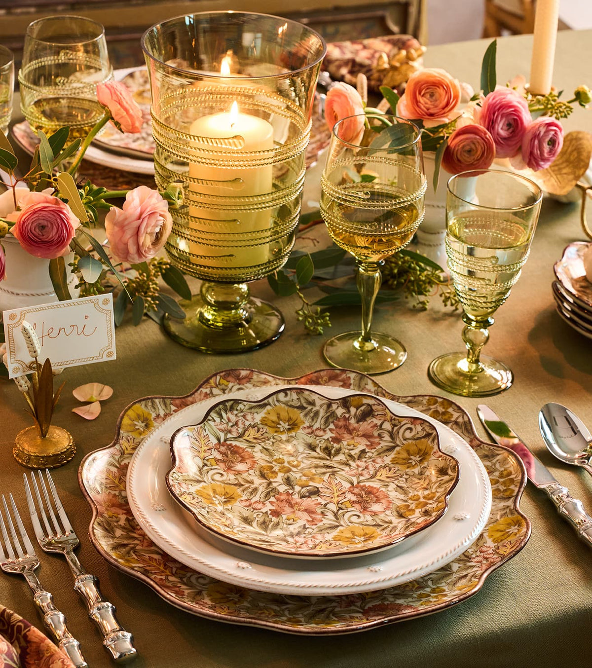 A beautifully set table for a special occasion with a floral patterned dinnerware set, a large lit candle in a glass hurricane vase, green-tinted wine glasses, and a floral arrangement.