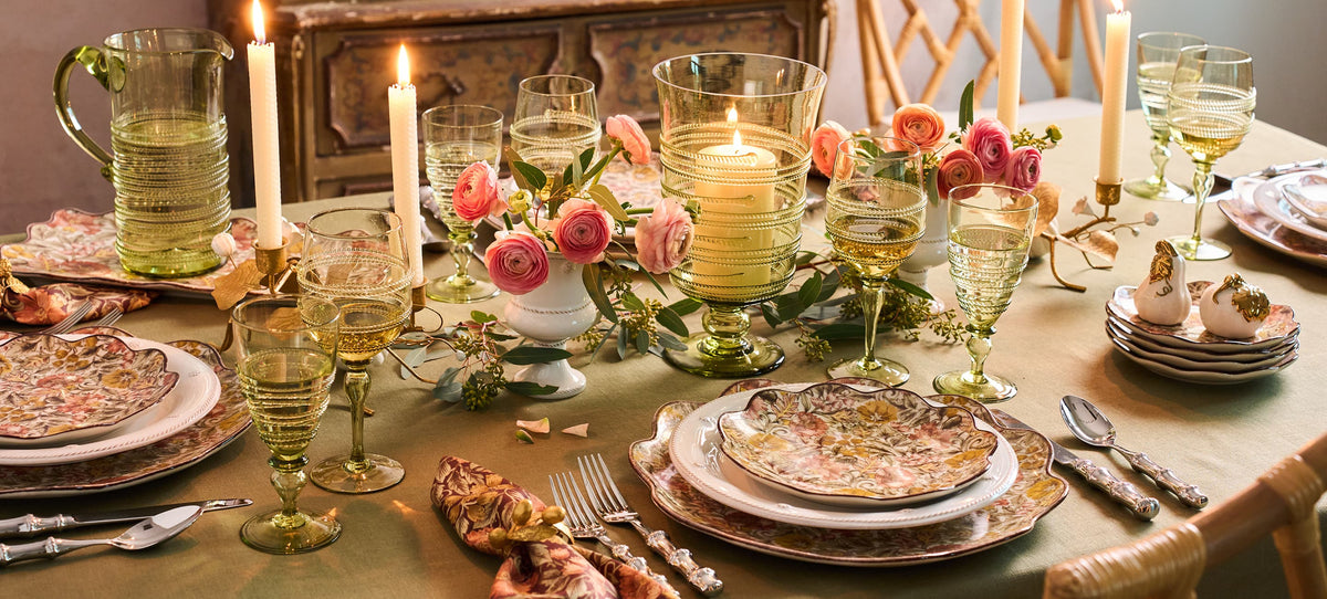 A styled dining table setting with vintage-style green glassware, layered plates, lit taper candles, and floral centerpieces in pink and peach roses.