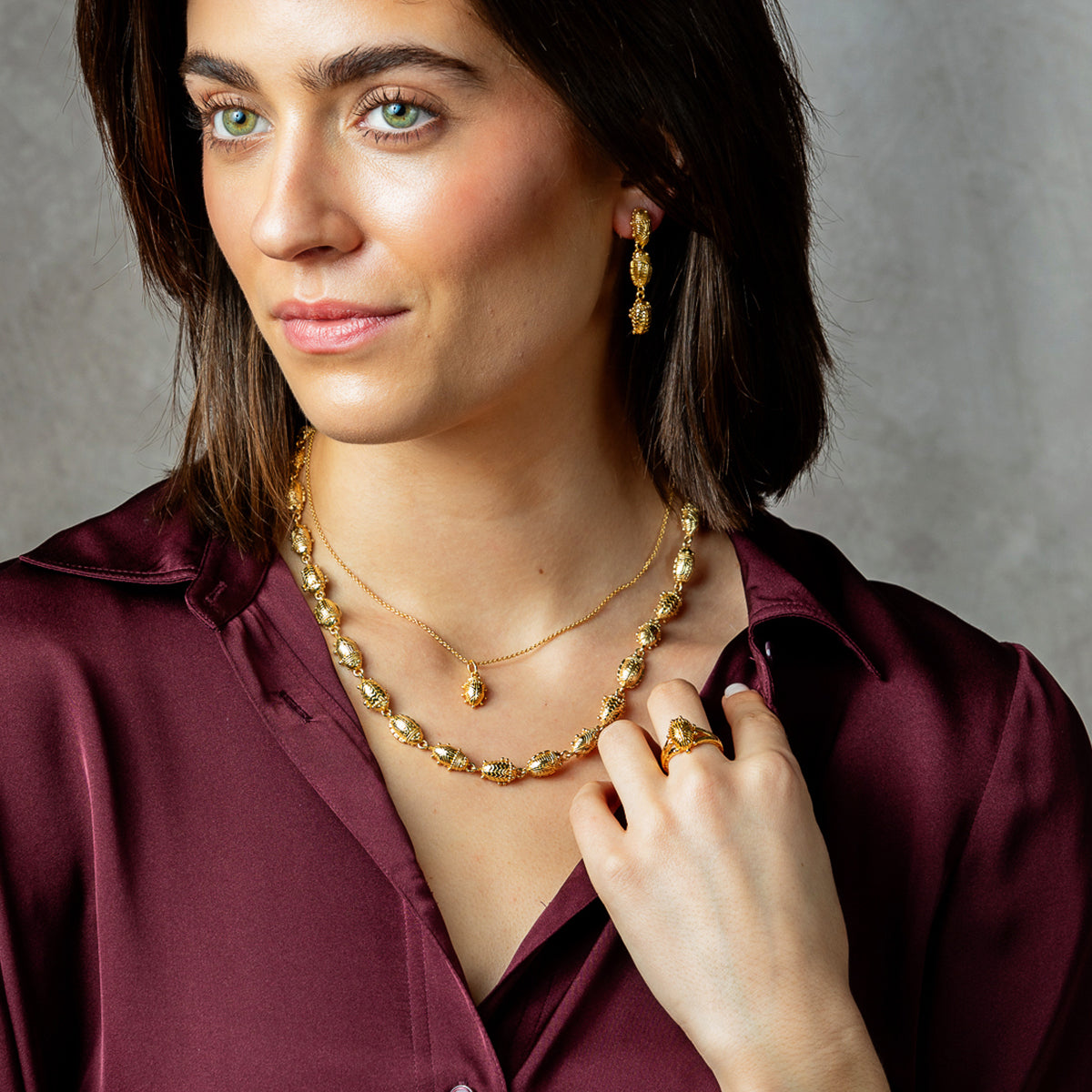 A woman wearing a gold beaded necklace and matching drop earrings, styled with a gold ring and a burgundy satin shirt against a textured gray background.
