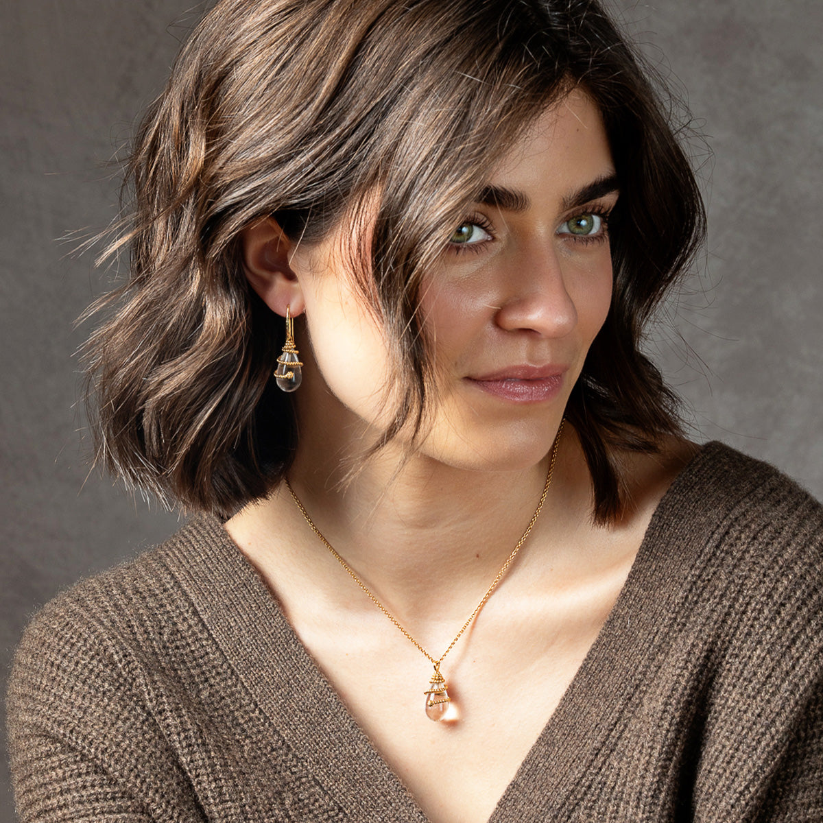 A woman with short brown hair wearing a delicate gold necklace and matching drop earrings with clear faceted gemstones, set against a neutral gray background.
