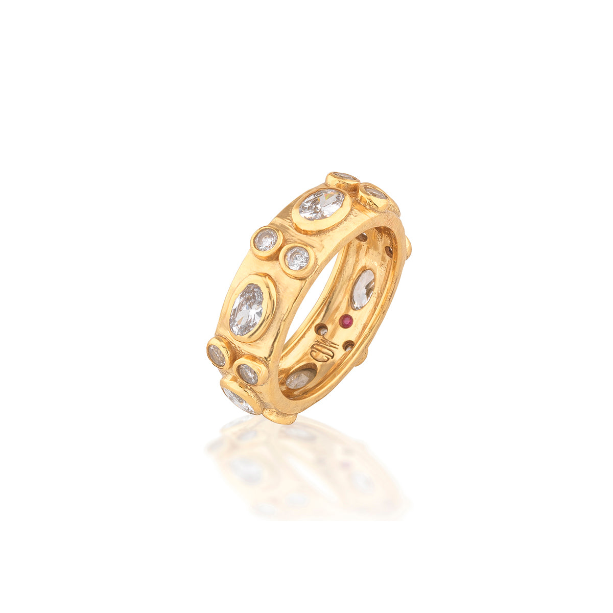 A gold band ring featuring multiple bezel-set clear gemstones and a small round red accent stone, shown on a clean white background with a soft reflection below.
