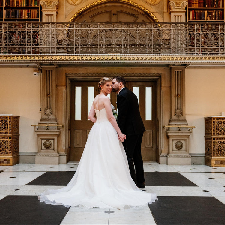 A bride in a white wedding gown and a groom in a black suit kiss in the grand interior of a library with ornate bookshelves and balconies.