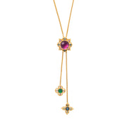 A gold lariat necklace featuring a large oval purple cabochon at the main pendant, with two dangling chains ending in floral and cross-shaped pendants set with green, blue, and small clear stones.