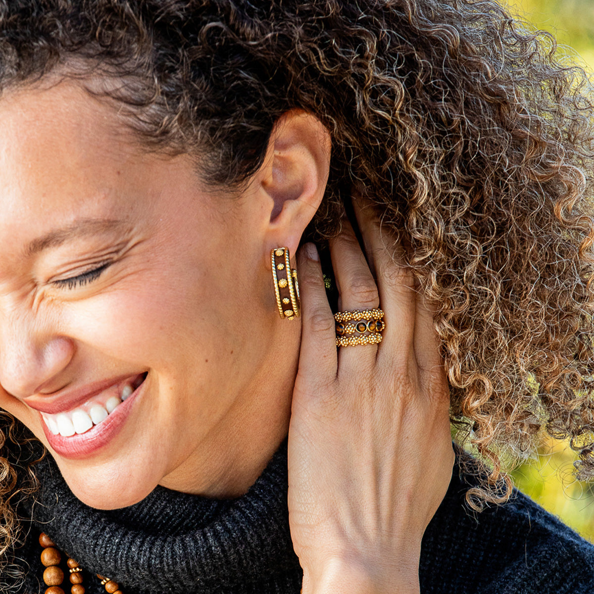 A close-up lifestyle shot of a smiling woman with curly brown hair wearing gold hoop earrings and matching gold rings with dark inlay details.