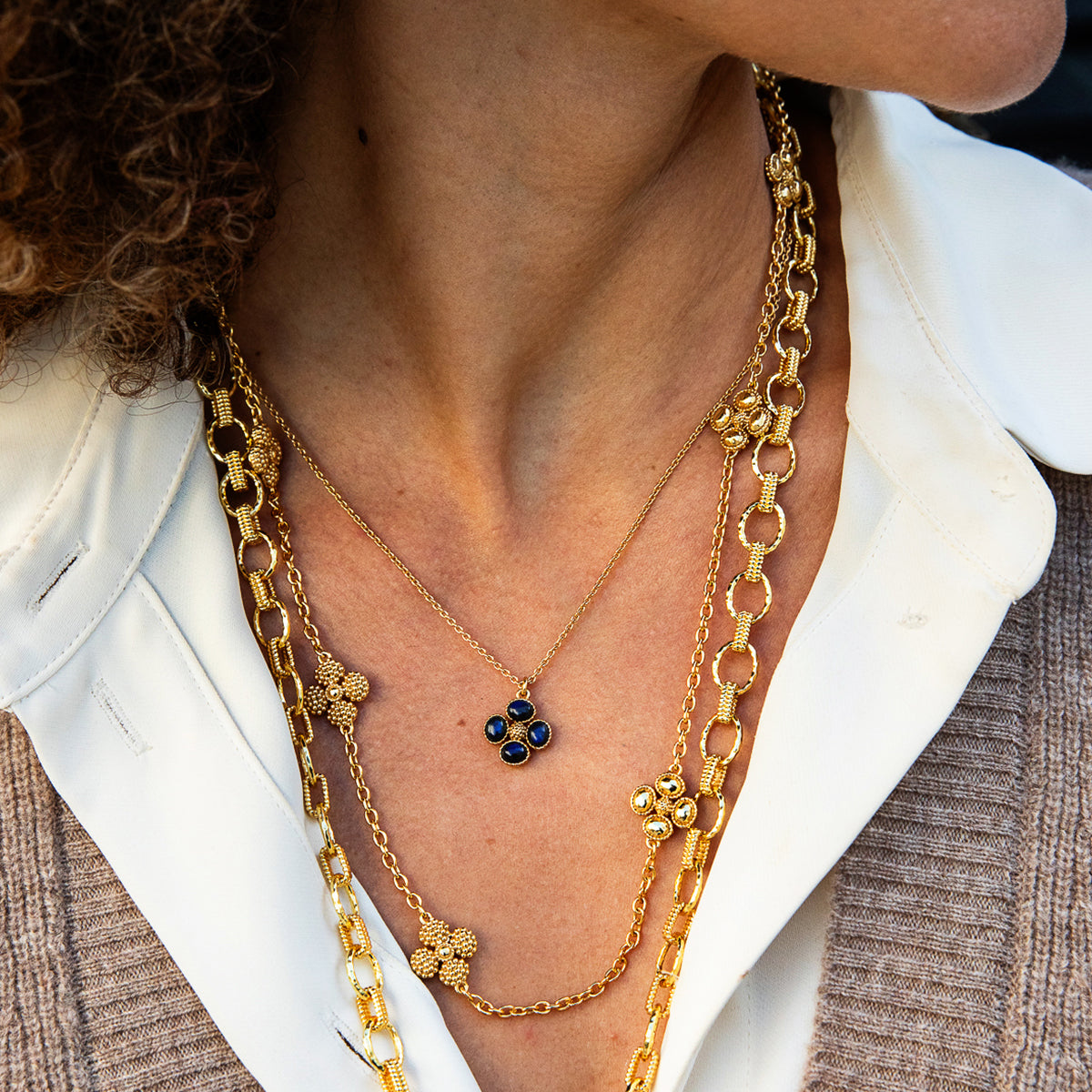 A close-up of a person wearing layered gold necklaces, including a delicate chain necklace with a dark blue floral clover pendant, layered over a white collared shirt.