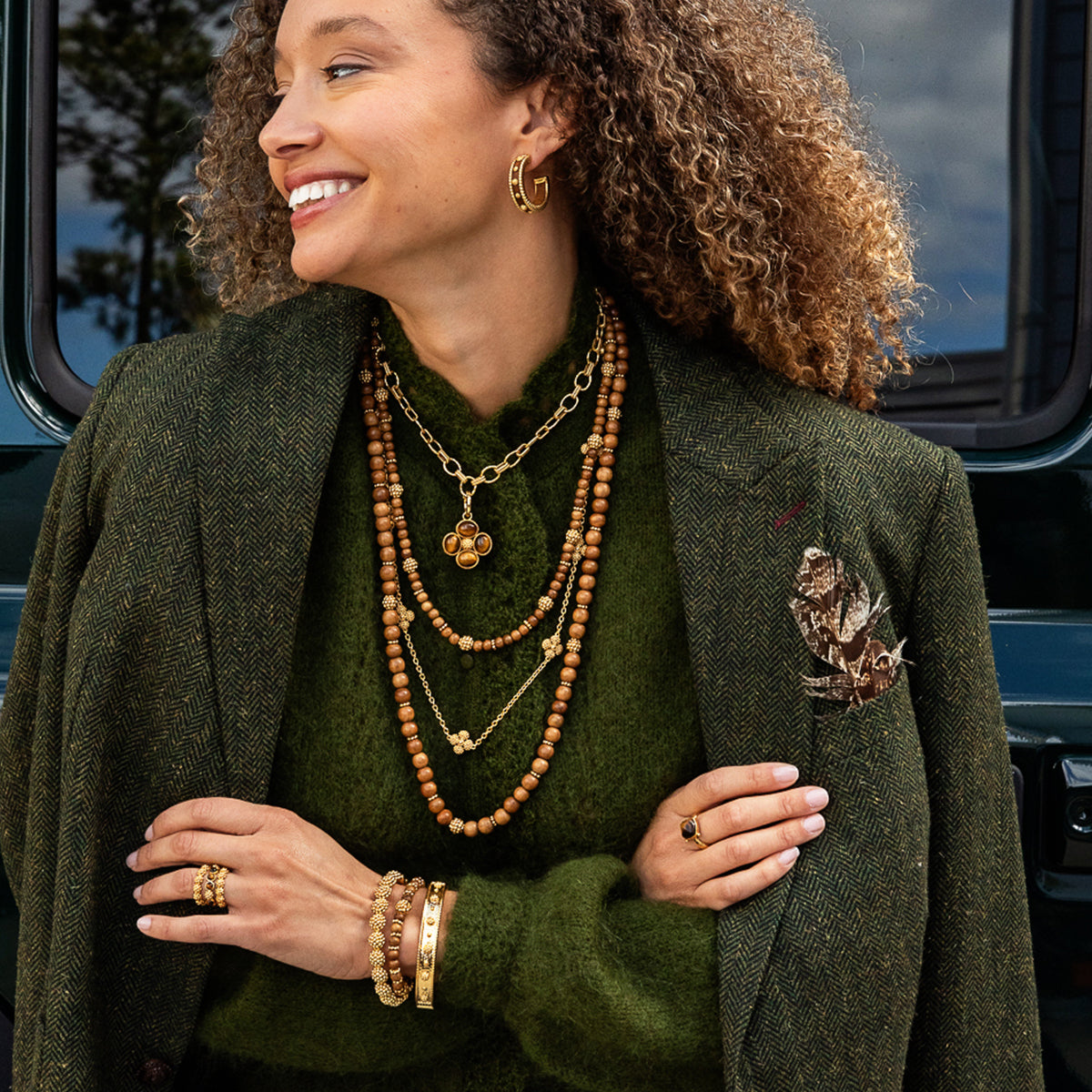 A woman wearing layered gold and wooden bead necklaces, rings, and earrings, styled with an olive green knit sweater and a textured dark green tweed blazer. She is posed outdoors in front of a dark vehicle window.