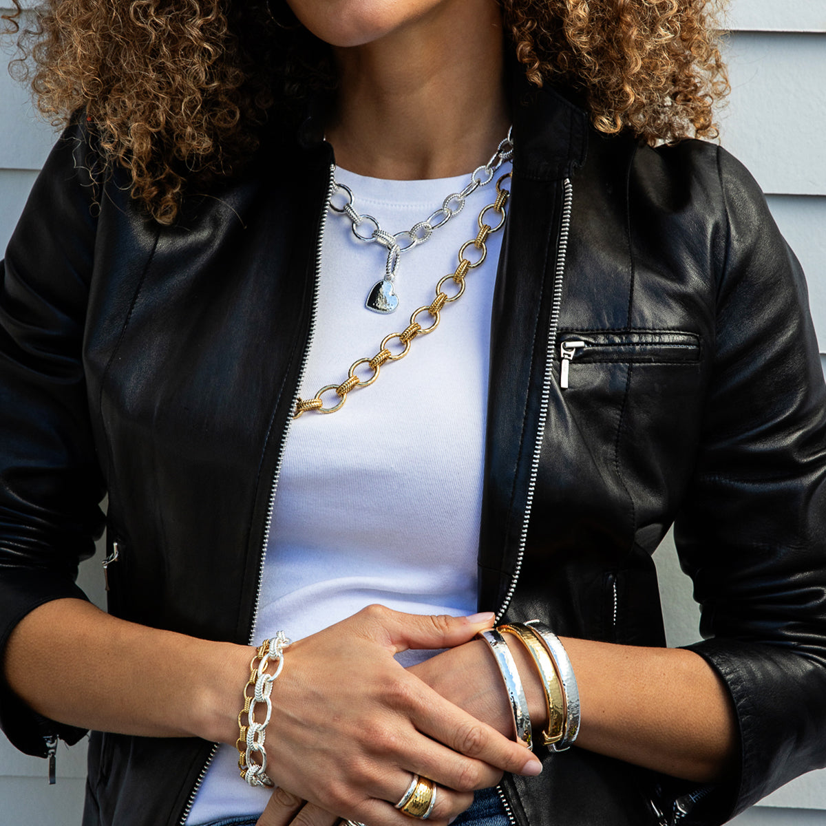 A close-up of a person wearing a black leather jacket over a white t-shirt, accessorized with layered gold and silver chain necklaces, bracelets, and rings.