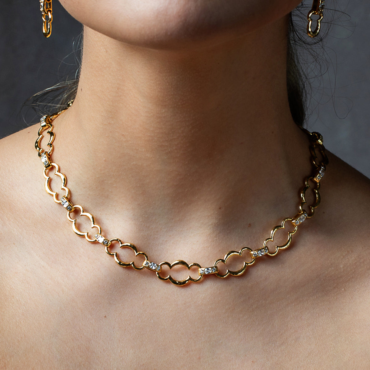 A close-up of a model's neck and upper chest wearing a gold chain necklace with interlocking oval links and small clear stones, with matching gold earrings partially visible.