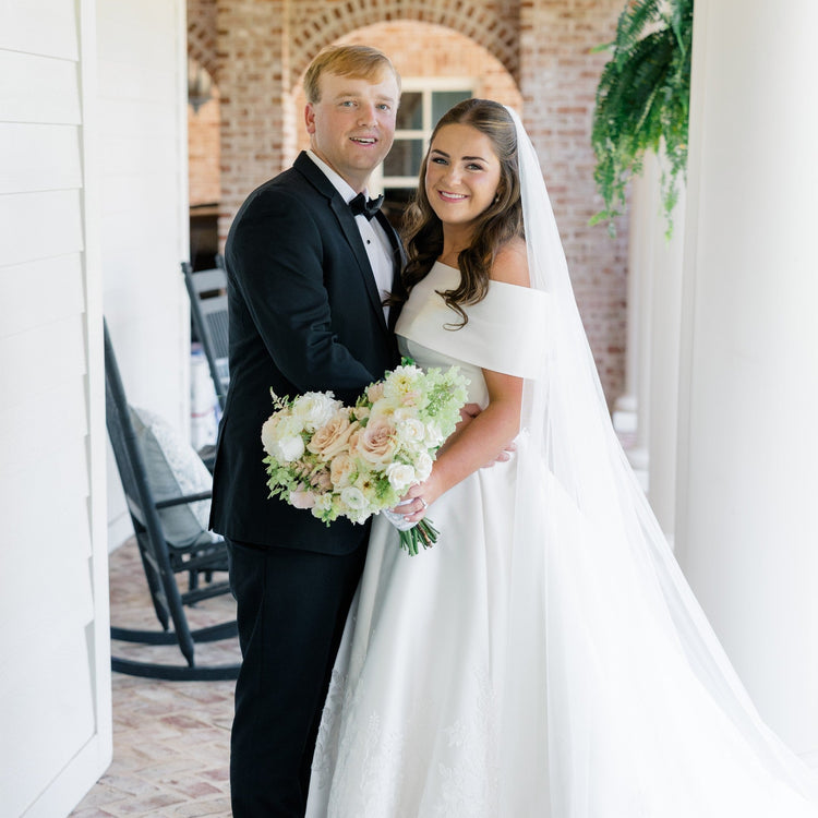 A bride in an off-the-shoulder white wedding gown and veil holding a bouquet of white and blush flowers, standing with a groom in a black tuxedo on a brick porch.