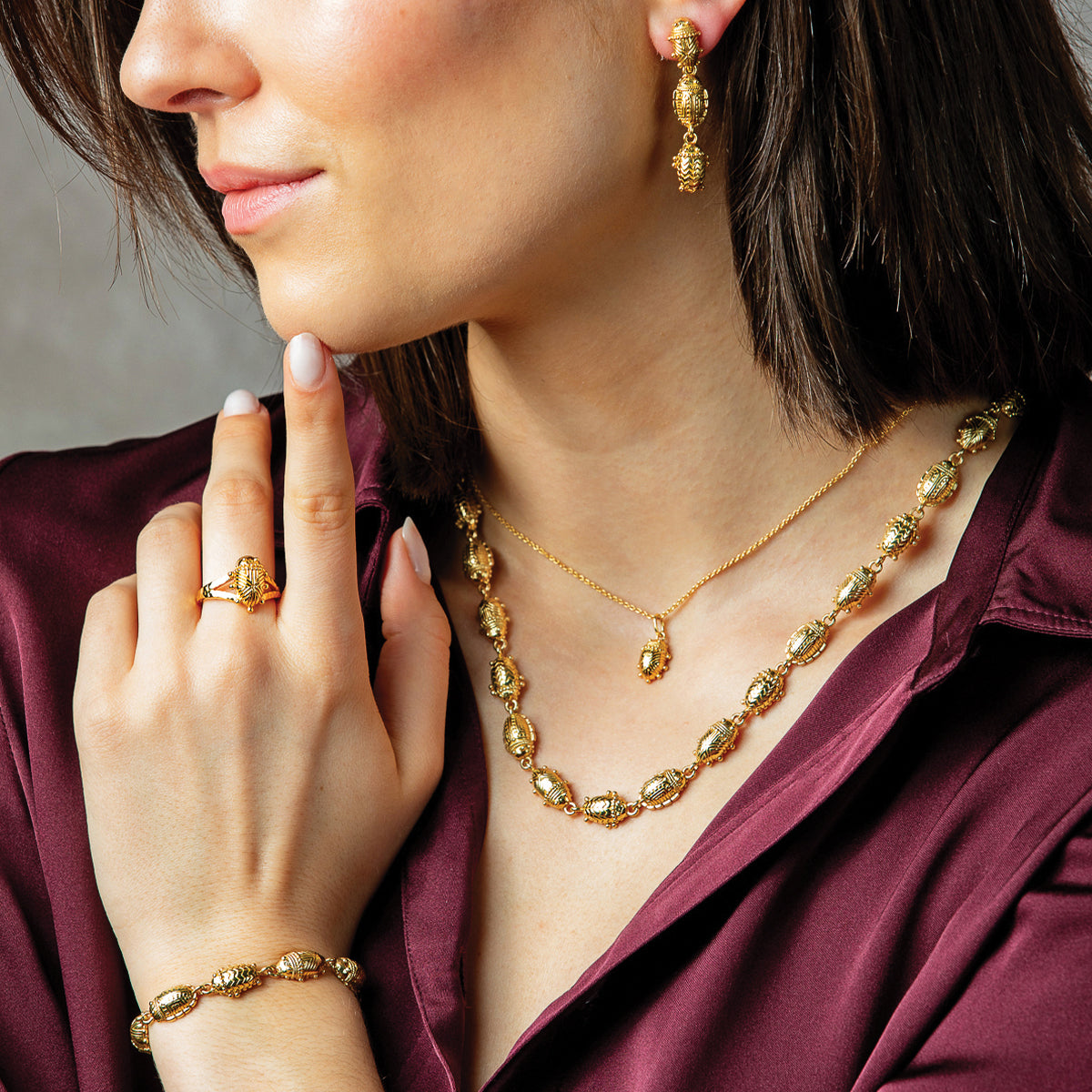 A close-up lifestyle shot of a woman wearing a matching gold jewelry set, including a necklace, ring, and bracelet, paired with a satin burgundy blouse.