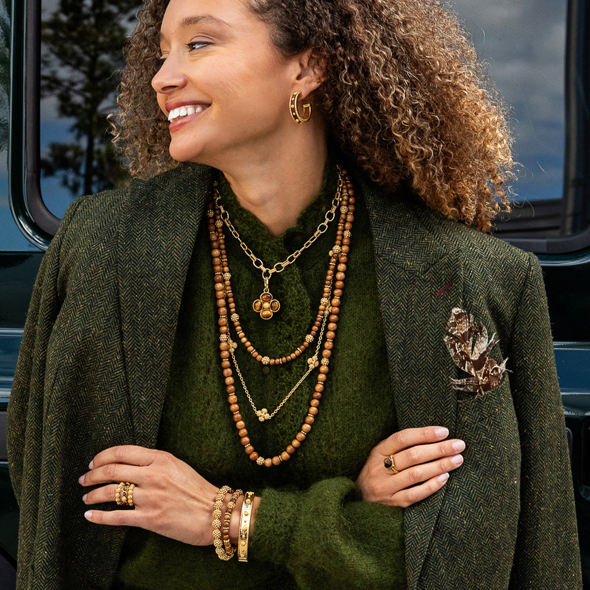 A woman with curly brown hair models layered gold jewelry, including a clover pendant necklace, beaded necklaces, hoop earrings, and rings, styled with an olive green sweater and tweed blazer.