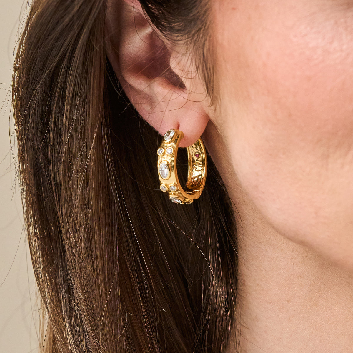 A close-up of a person's ear wearing a small gold hoop earring with small clear stones set along the front edge, against a soft beige background.
