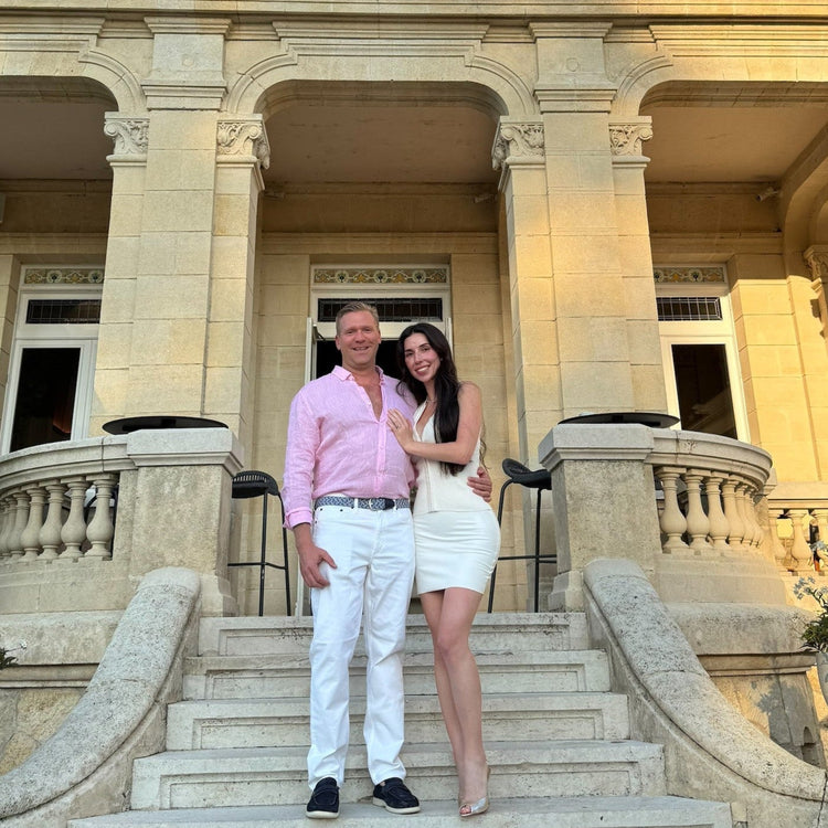 A man and a woman posing together on the stone steps of a grand, elegant building with classical columns and dormer windows under a blue sky.