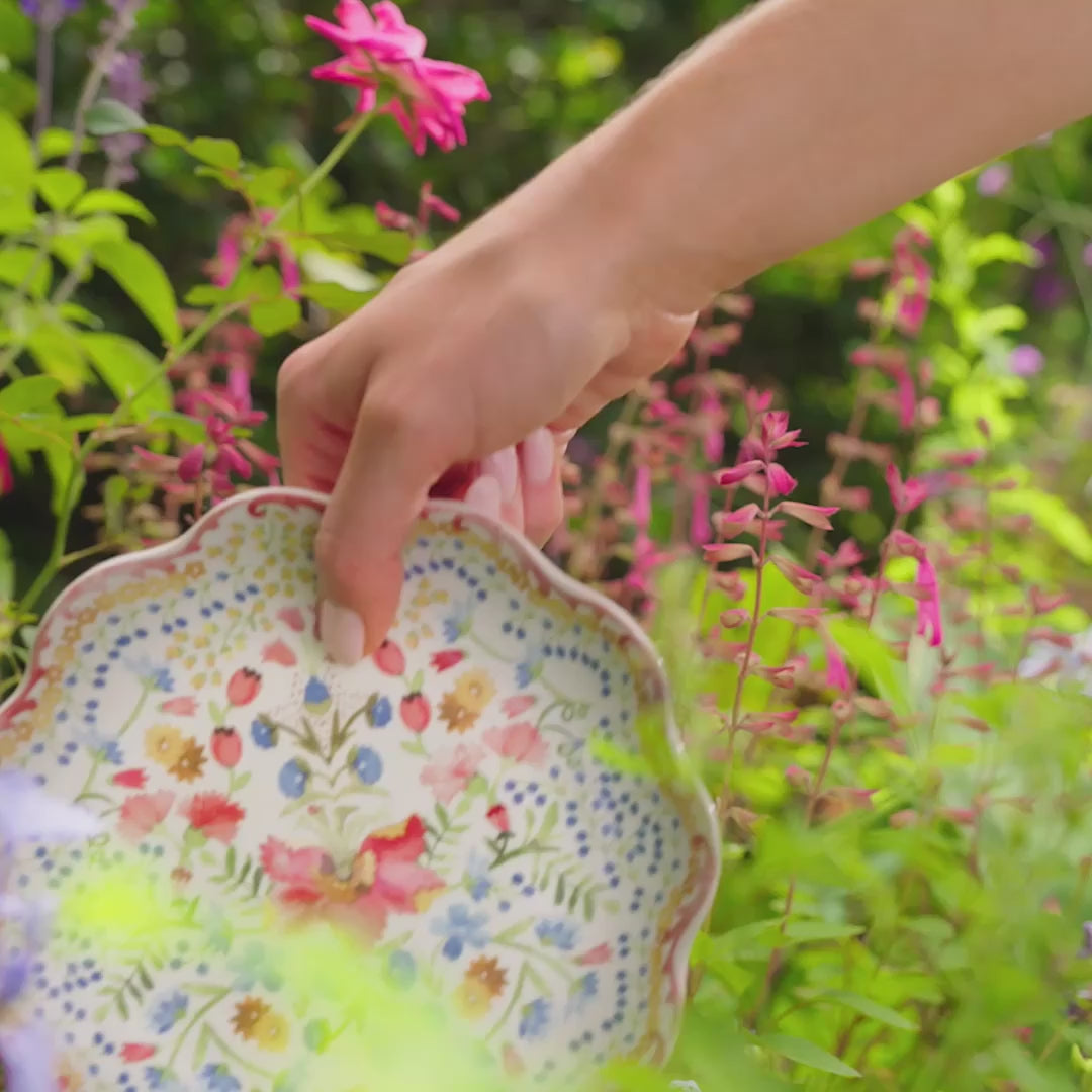Load video: Video featuring a woman setting up a floral tablescape with floral plates, candlesticks, and place card holders in a garden.