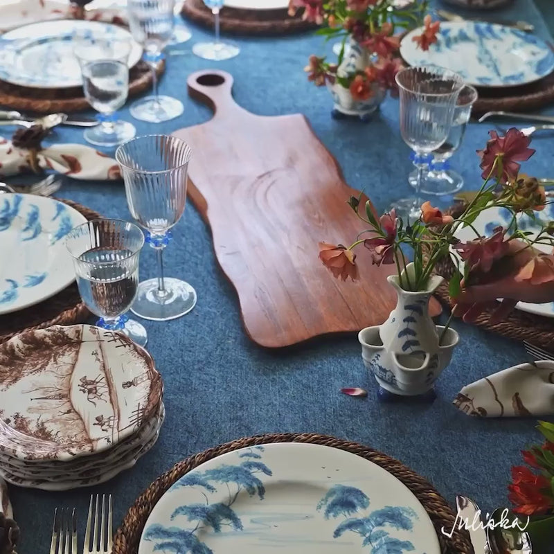 Load video: Moveable video featuring woman's hand setting and arranging the tablescape. The tablecloth is denim blue, the plates are brown and blue, and the glasses are clear with blue accents.