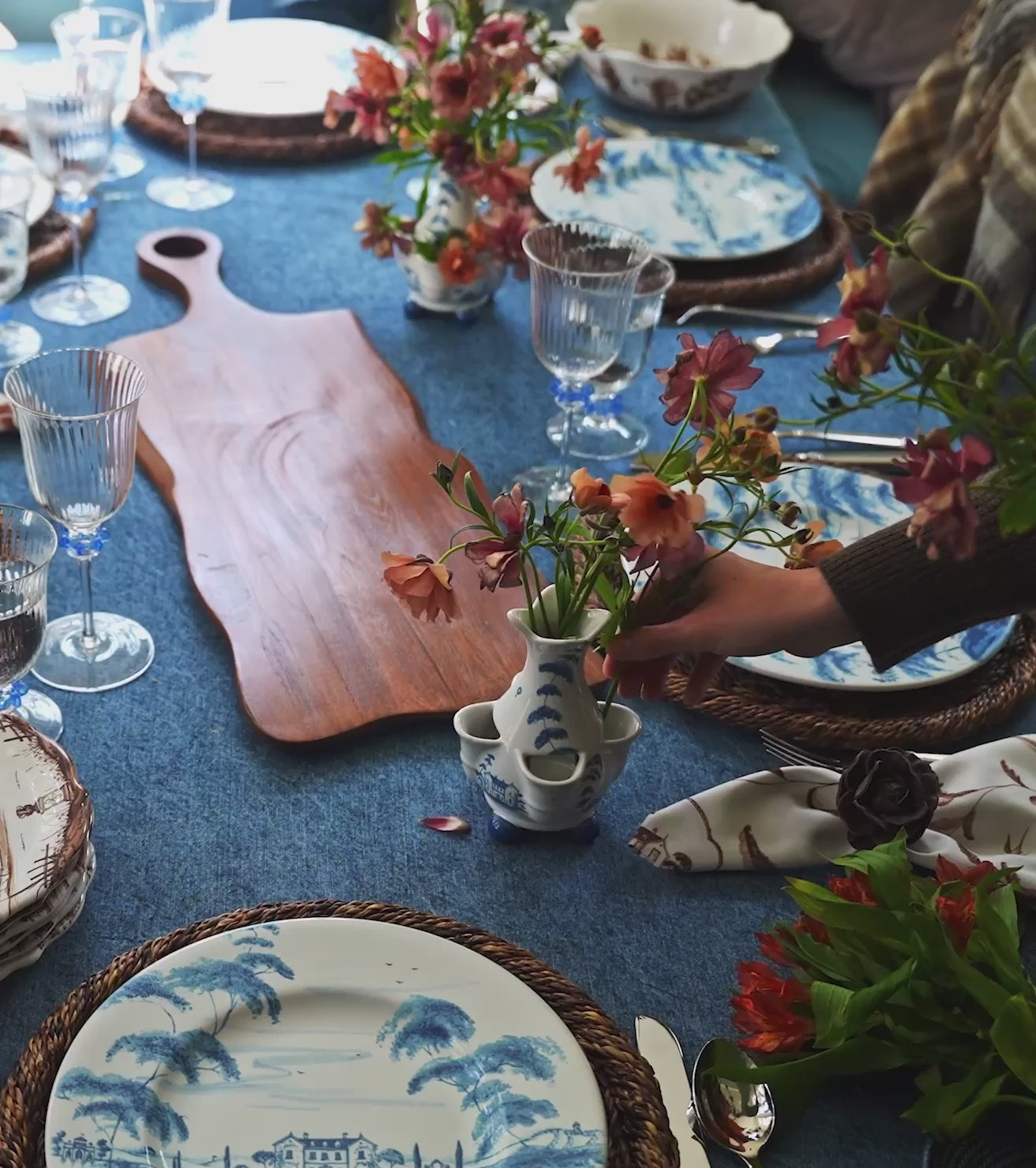 Load video: Moveable video featuring woman's hand setting and arranging the tablescape. The tablecloth is denim blue, the plates are brown and blue, and the glasses are clear with blue accents.